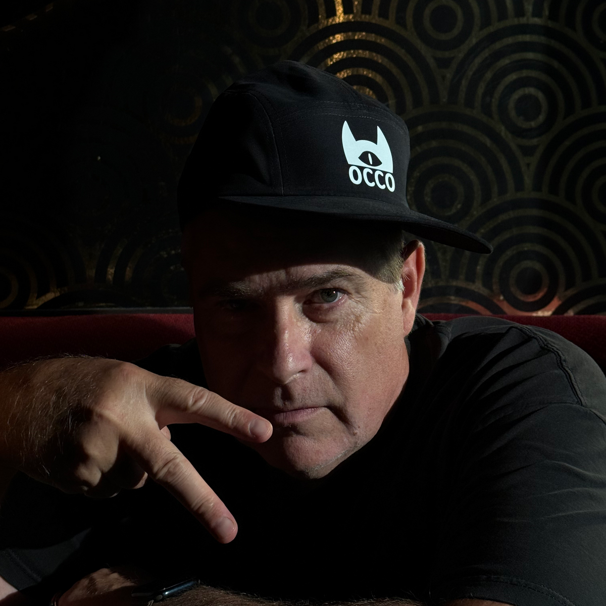 A man wearing a black baseball cap with a white 'OCCO' logo, resting his head on a red couch against a dark patterned wall.